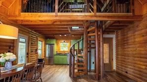 A log cabin interior with a dining area, kitchen, and a staircase leading to a loft.