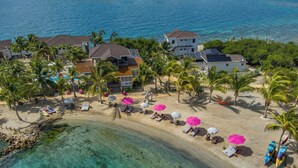 An aerial view of a beachfront property with multiple houses, a sandy beach with lounge chairs and umbrellas, and a clear blue ocean. - Magenta Caye Luxury Private Island Resort - All inclusive (Placencia)
