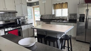 A modern kitchen with white cabinets, a stainless steel refrigerator, a large island with stools, and a window with curtains.