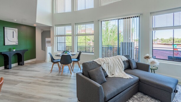 Un salon moderne avec un canapé gris, un coin repas avec des chaises et de grandes fenêtres.