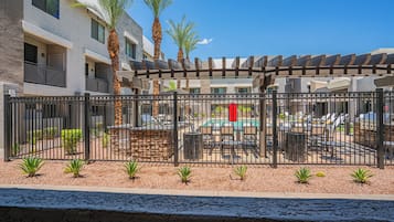 A pool area with lounge chairs and a pergola, surrounded by a fence and palm trees.