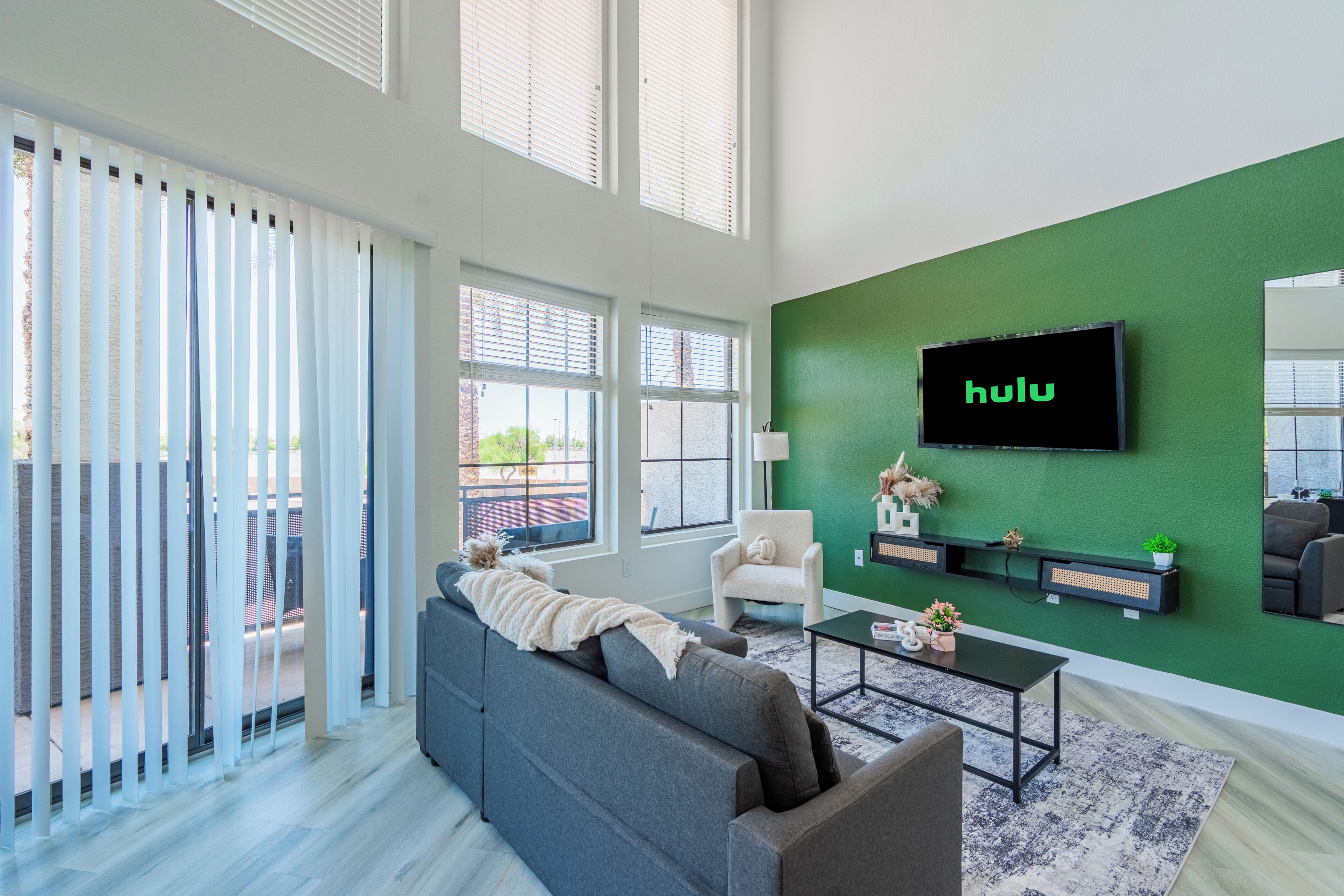 A modern living room with a grey sofa, a coffee table, a green accent wall, and a TV mounted on the wall displaying 'hulu'.