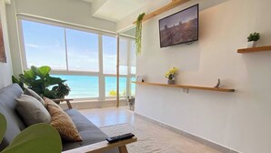 Ett modernt vardagsrum med ett stort fönster med utsikt över havet, en grå soffa, trähyllor och en väggmonterad TV.
