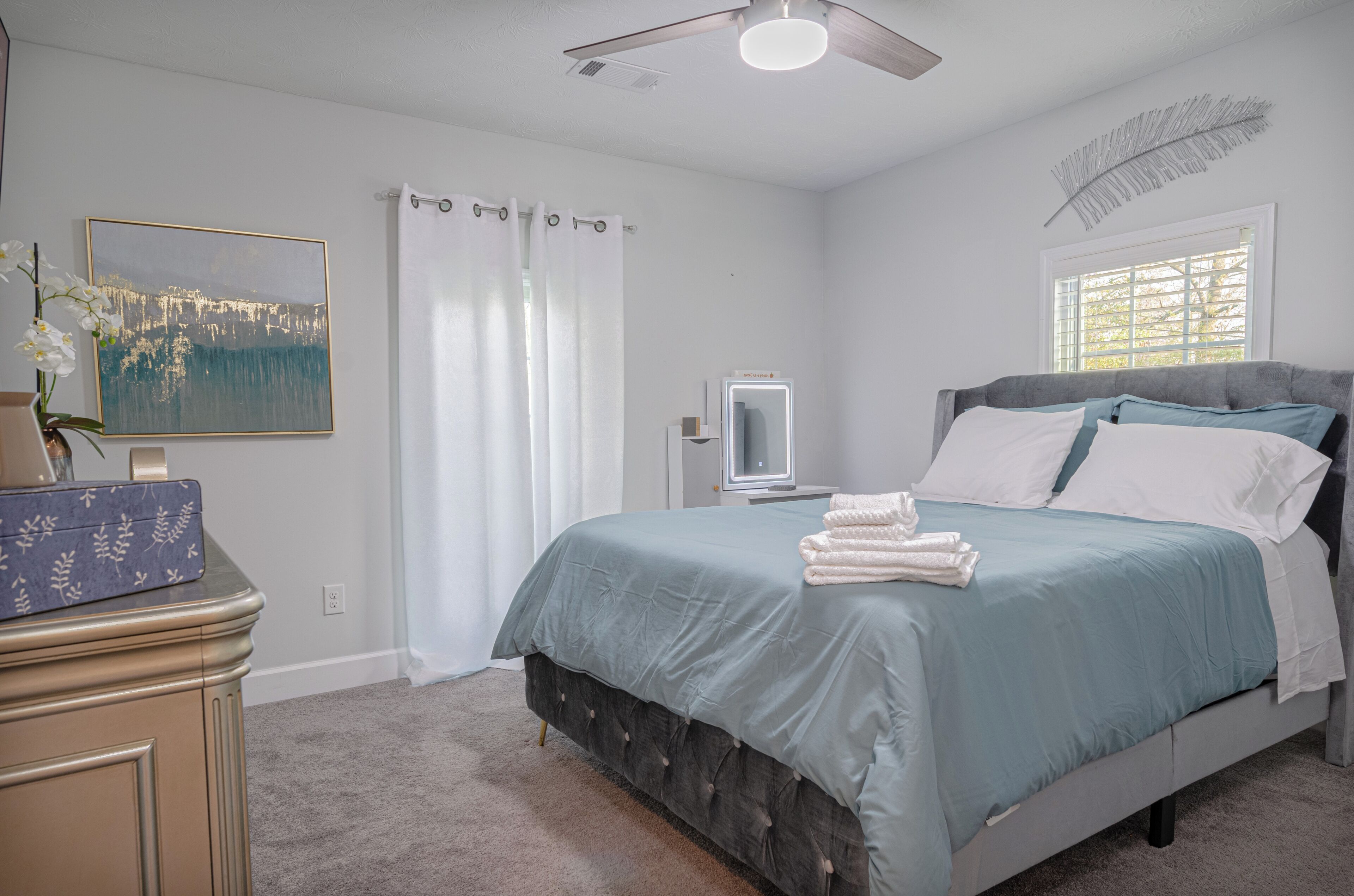 A bedroom with a bed, a vanity, a mirror, a ceiling fan, and a window with curtains.