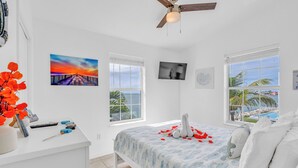 Ein Schlafzimmer mit Bett, Schreibtisch, Fernseher und großen Fenstern mit Blick auf einen Pool und Palmen.