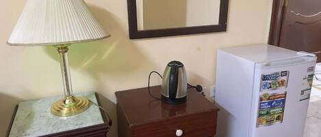 Una habitación pequeña con una mesita de noche de madera, una lámpara, un hervidor y un refrigerador.