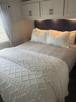 Ein ordentlich bezogenes Bett mit einer gemusterten Tagesdecke und zwei Kissen, ein dunkles Kopfteil und weiße Schränke darüber.