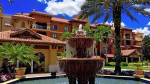 A multi-tiered fountain in a courtyard with a pool, surrounded by palm trees and buildings with Mediterranean-style architecture. - Christmas at Disney! Spacious 2BR Condo w/ Lazy Rivers, Pools, & Fireworks View (Lake Buena Vista)