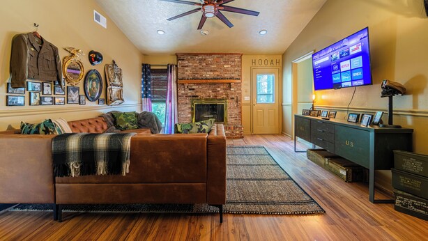 A living room with a brown sofa, a ceiling fan, a brick fireplace, and a mounted TV. - Fort Winburn Military Home (Columbus)