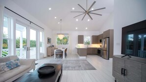 A modern living room with a sofa, a coffee table, and a TV. There is a dining area with a table and chairs. The kitchen has a refrigerator, stove, and cabinets. - Brand New Designer Home on Banks Road, Steps from Beach, Walk to La Bougainvillea (Governor's Harbour)