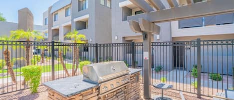 Un patio con una parrilla de piedra, un pérgola y un acceso cerrado a un edificio residencial.