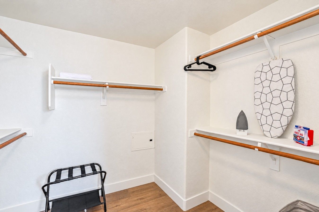 A room with a wooden floor, white walls, and a black metal chair. There are two wooden beams on the wall, one with a white towel hanging and another with a black hanger. A white shelf with a hexagonal mirror and a small iron is also visible.