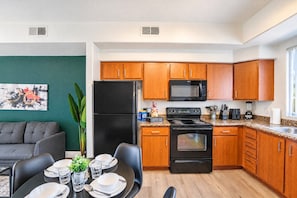 A modern kitchen with wooden cabinets, a black refrigerator, a stove, and a microwave. There is a dining area with a table set for two. - Walk To Westgate w/ Modern Pool, Gym, Parking, W/D (Glendale)