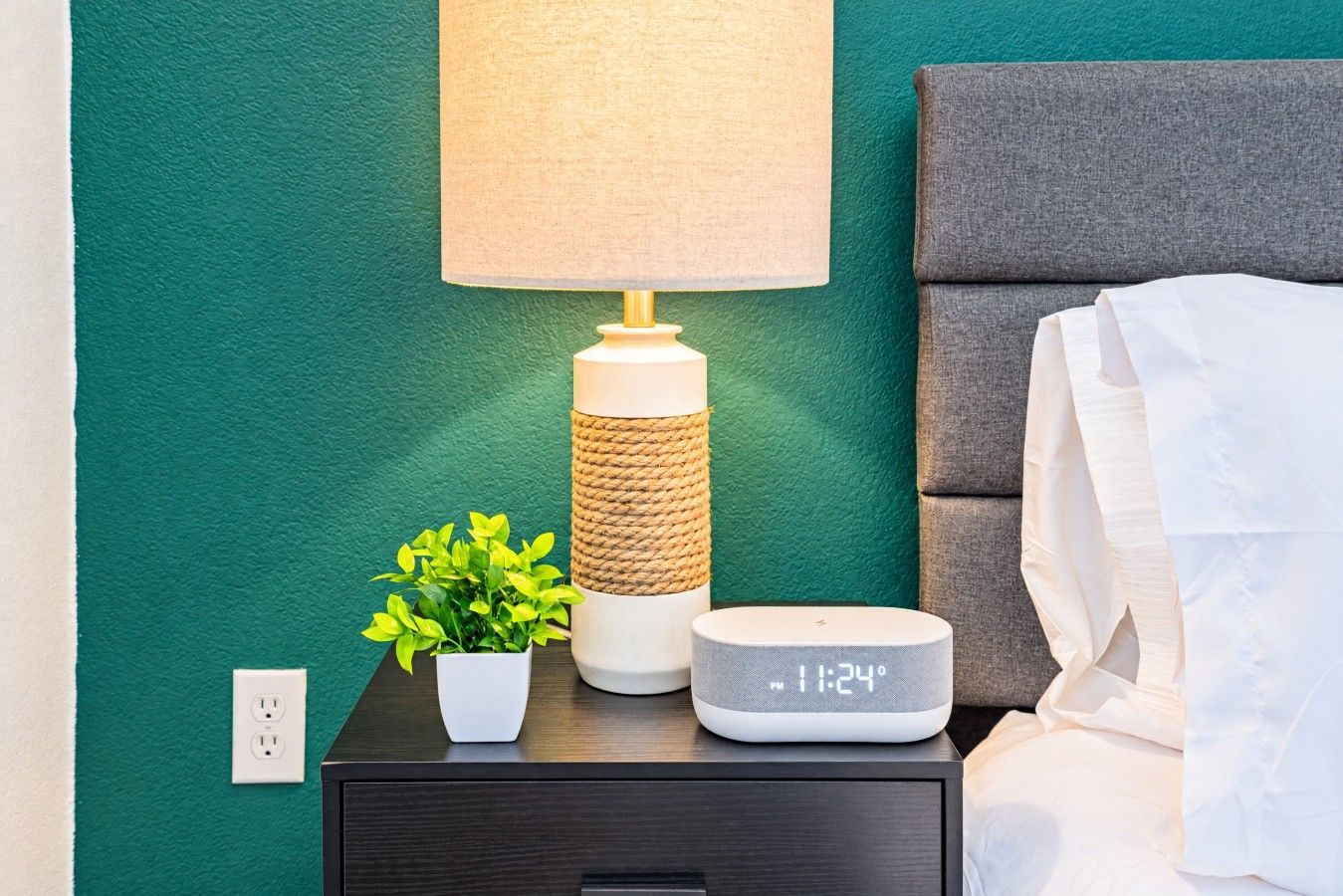 A bedside table with a lamp, a digital clock, and a small potted plant.