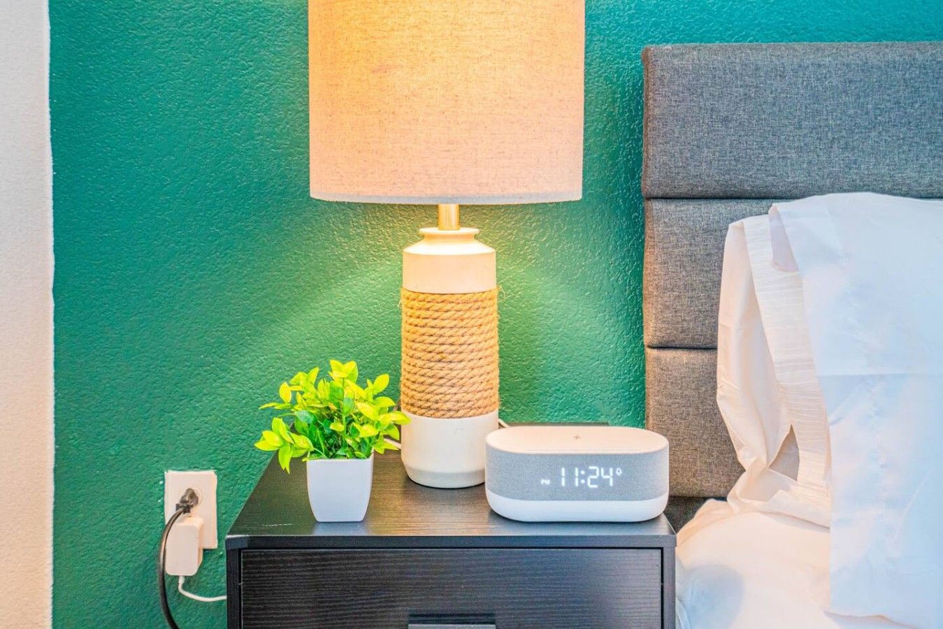 Una mesita de noche con una lámpara, un reloj digital y una planta en maceta.