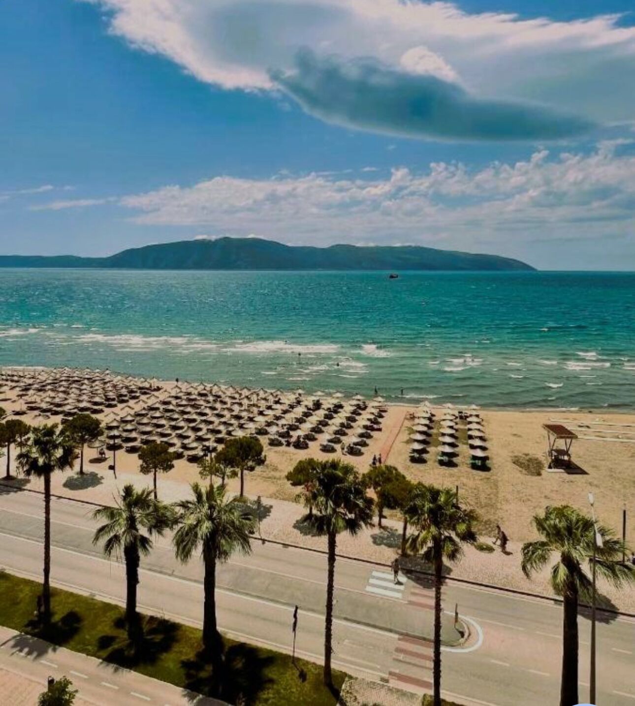 Une plage bordée de rangées de parasols et de palmiers, une route au premier plan et une étendue d’eau avec des montagnes en arrière-plan.