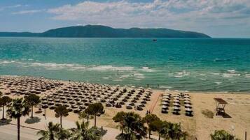 Une plage bordée de rangées de parasols et de palmiers, une route au premier plan et une étendue d’eau avec des montagnes en arrière-plan.