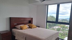 A bedroom with a large window, a wooden bed, and a bedside table.