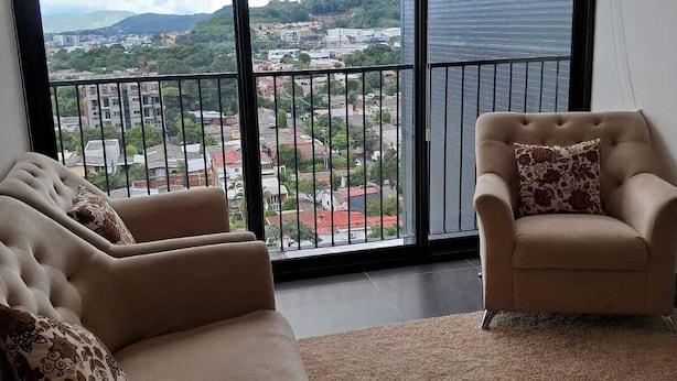A living room with a sofa and armchair, a large window with a view of a cityscape and hills.