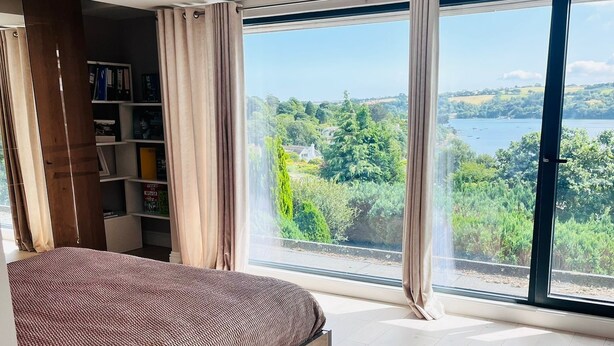 A modern bedroom with a large bed, a built-in bookshelf, and a view of a lake and greenery. - Spacious Modern Creek View House (Truro)