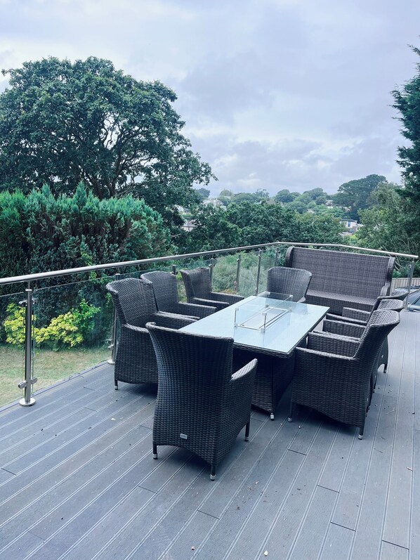 An outdoor patio with wicker furniture and a glass table, surrounded by greenery and overlooking a residential area.