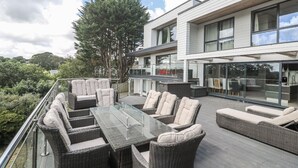 A modern outdoor patio with wicker furniture and a glass table, overlooking a lush garden and a contemporary building. - Spacious Modern Creek View House (Truro)