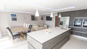 A modern kitchen with a central island, a dining area with a wooden table, and a sofa. - Spacious Modern Creek View House (Truro)