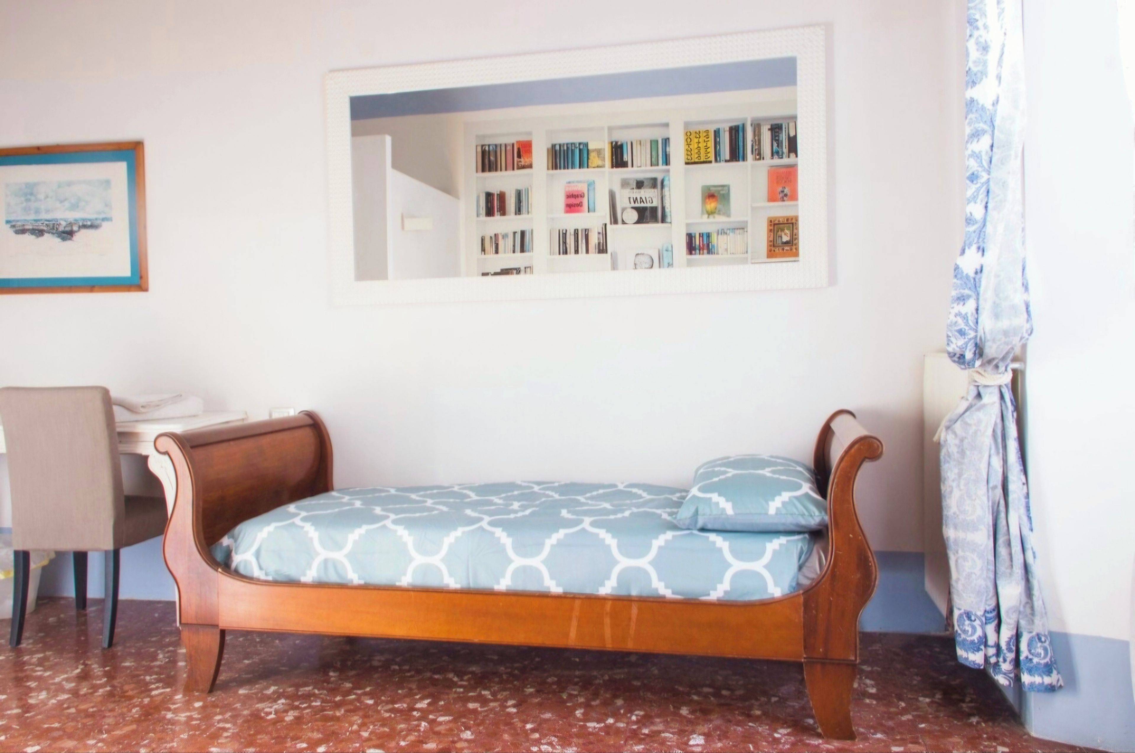 Un lit simple avec un matelas à motifs, une tête de lit en bois, un bureau avec une chaise, une bibliothèque et un tableau encadré accroché au mur.