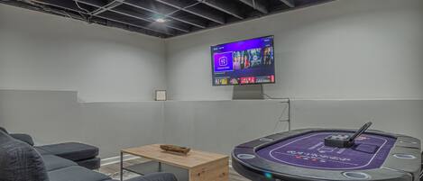 Een kamer met een wandgemonteerde televisie, een klein houten tafeltje, een poker tafel en grijze banken.