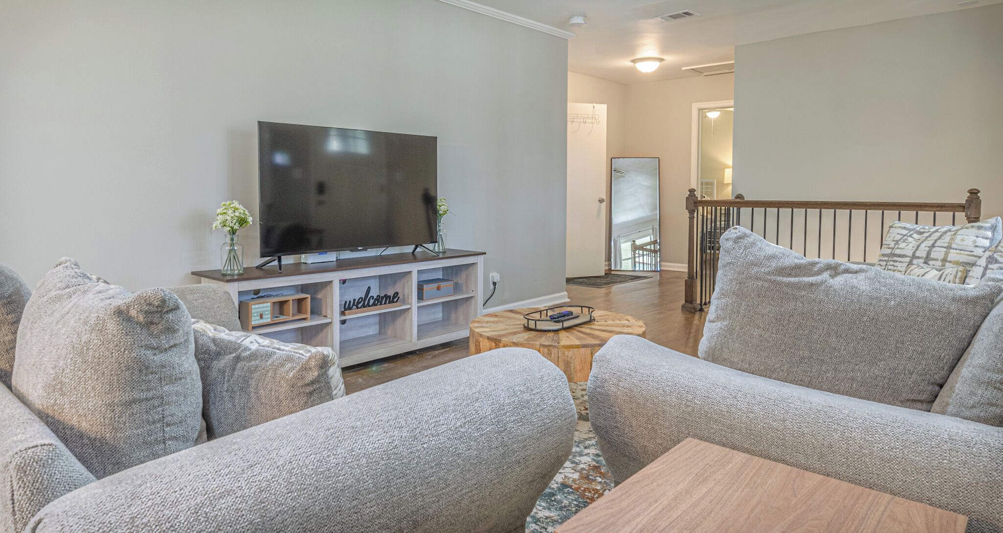 A modern living room with a flat-screen TV on a wooden stand, grey upholstered sofas, a coffee table, and a 'welcome' sign on the entertainment unit.