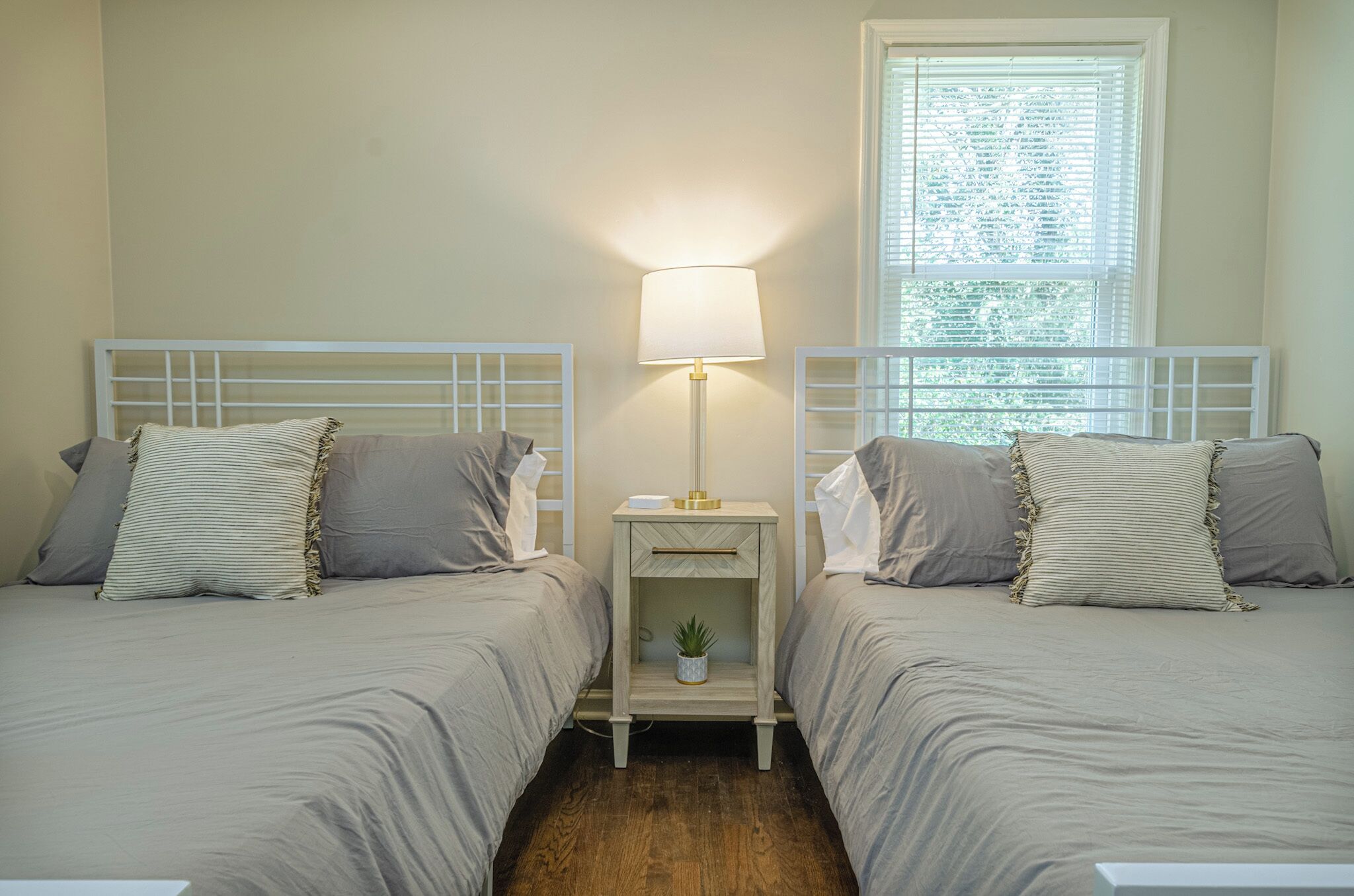 A room with two beds, each with a headboard, a bedside table with a lamp, and a window with blinds.
