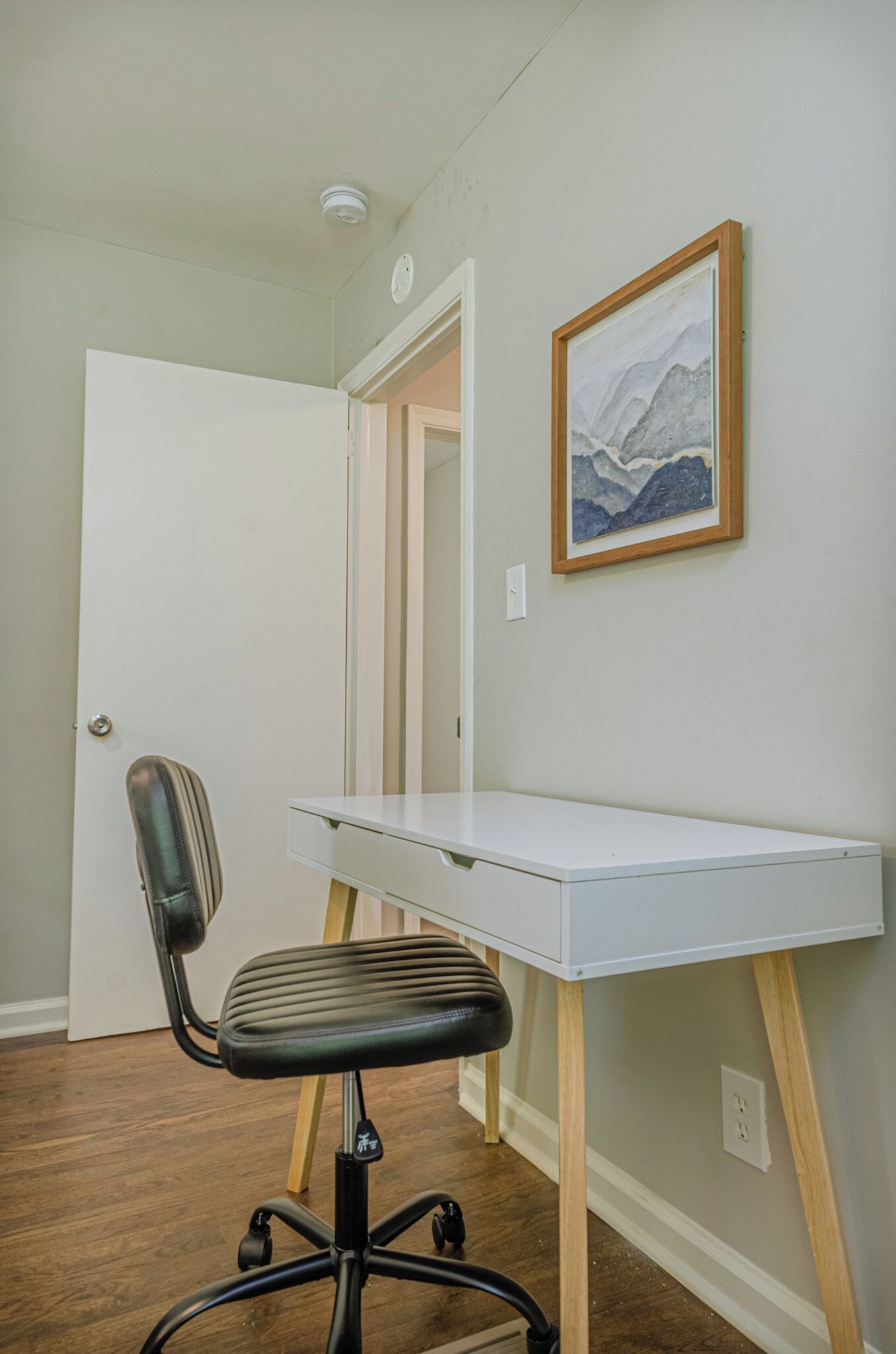 A room with a white desk, a black office chair, a framed picture on the wall, and a door leading to another room.