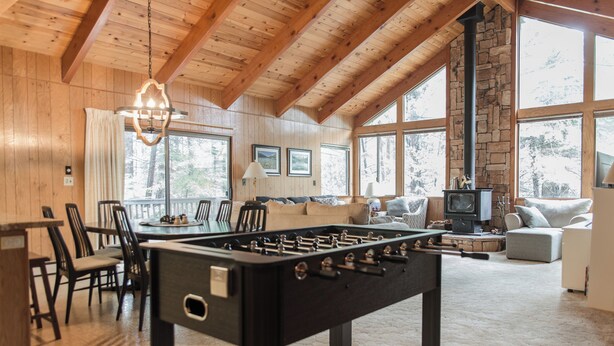 A cozy room with a foosball table, a stone fireplace, and large windows offering a view of the outdoors. - Amazing 3-bedroom Home in Beautiful Blue Lake Springs, Arnold California (Arnold)