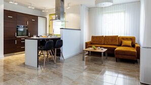 A modern kitchen with a central island, dark wooden cabinets, and stainless steel appliances. There is a sofa and a coffee table in the living area, and a large window with sheer curtains.