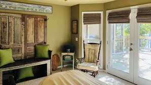 A room with a large wooden bench, a chair, and a bed. There is a door leading to a balcony with a view of the outdoors.