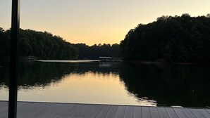 A wooden deck overlooking a calm lake with a forested shoreline and a small dock in the distance.