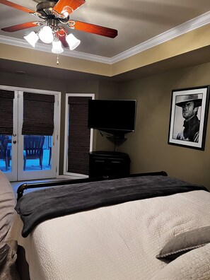 Un dormitorio con cama, ventilador de techo, televisor y una puerta con persianas.