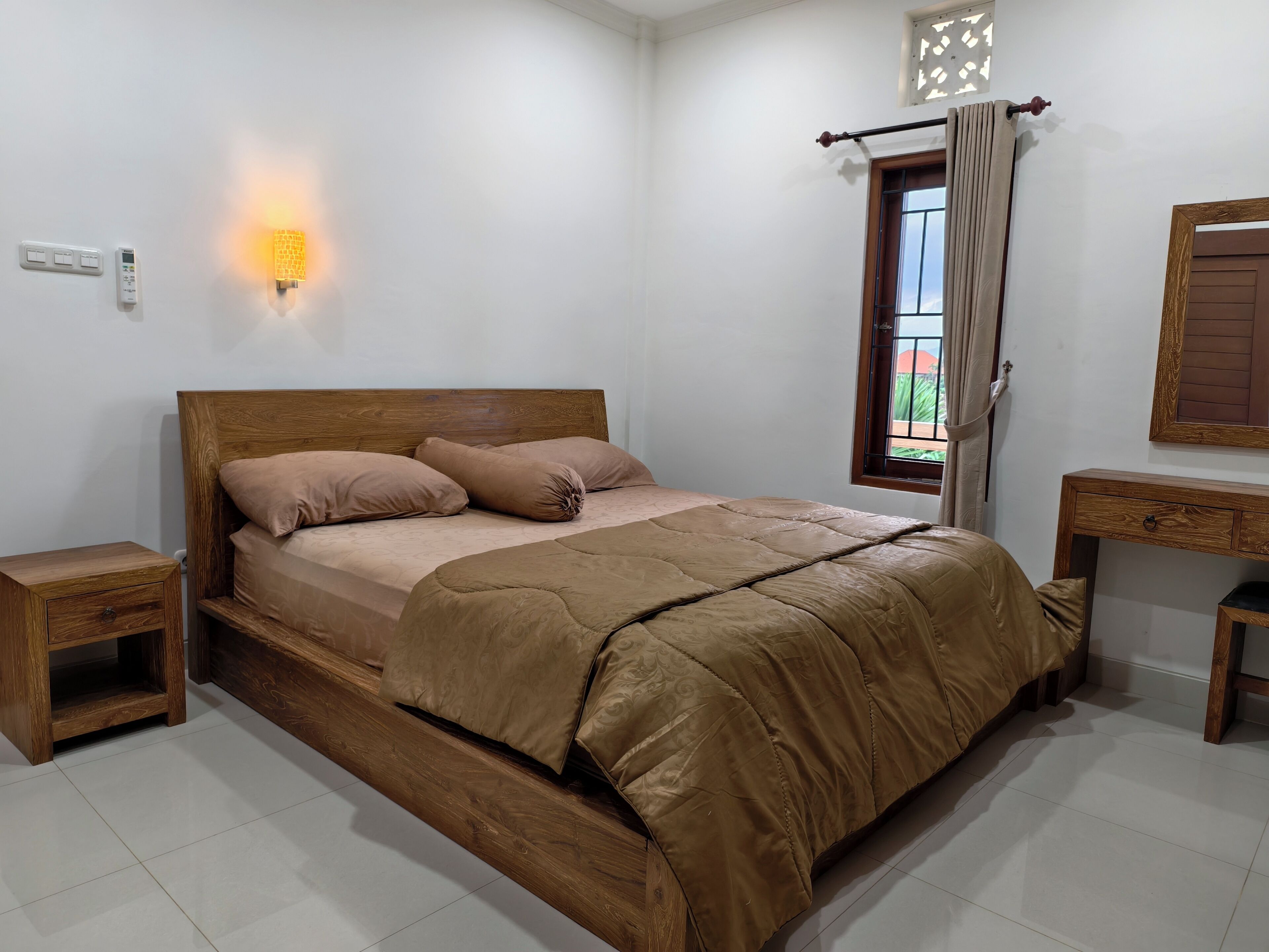 Un dormitorio con cama de madera, una mesita de noche, una lámpara de pared, una ventana con cortinas y un escritorio de madera.