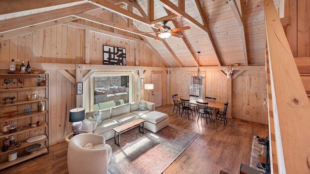 A cozy loft-style room with a high ceiling, wooden beams, a large window, a sofa, a dining table, and a chandelier. - Cozy Mountain Cabin w/ Creek, near Dining & Hiking (Monteagle)