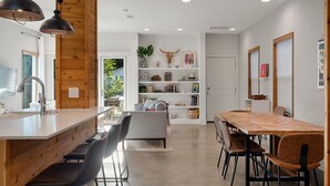 A modern kitchen with a wooden accent wall, a central island with bar stools, a dining area with a long wooden table and chairs, and a built-in bookshelf with decorative items. - Group-Friendly 4BR | Steps from East Austin Fun (Austin)