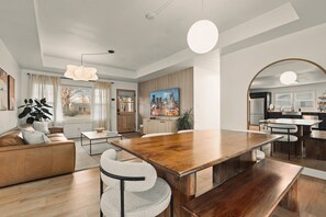 A modern living room with a wooden dining table, a sofa, and a large window with curtains.