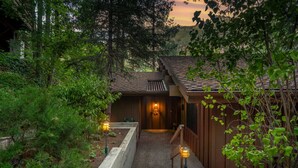 A pathway leading to a wooden building with a sloped roof, surrounded by dense greenery.