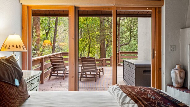 A bedroom with a large bed, a nightstand, a lamp, and a view of a deck with chairs and a grill.