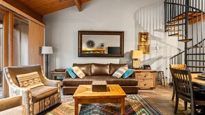A cozy living room with a brown leather sofa, a wooden coffee table, and patterned rugs. A spiral staircase is visible in the background.