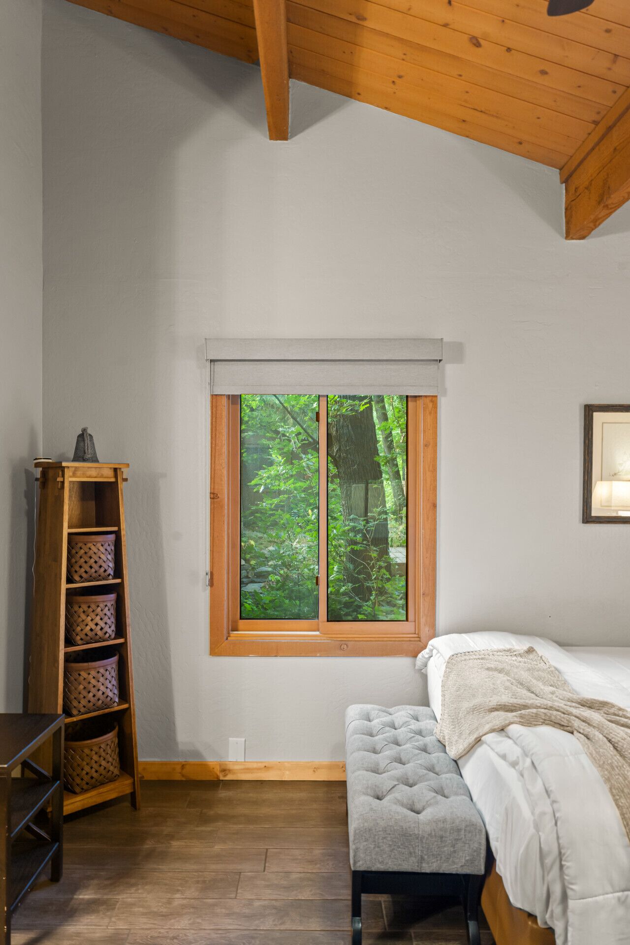 A bedroom with a wooden ceiling, a window with a view of greenery, a bed with a white comforter, and a wooden shelf with baskets.
