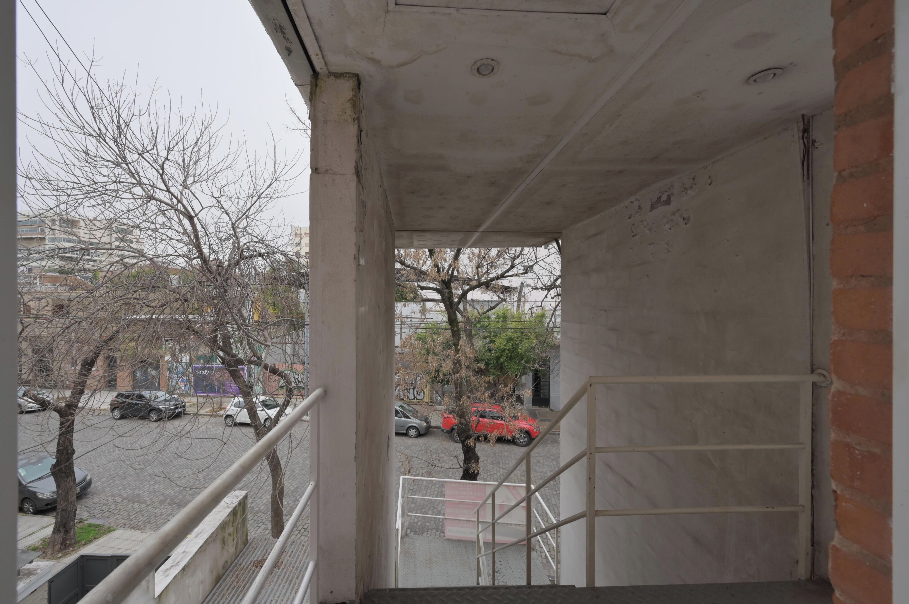 A view from a balcony overlooking a parking area with several cars, including a red car, and leafless trees.