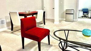 A modern living room with a glass coffee table, a red chair, a dining table with chairs, a small refrigerator, and a computer desk. - Entire basement with large windows, private full bathroom and office (Ottawa)