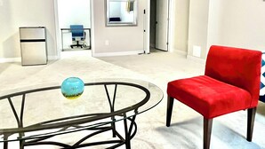 A modern living room with a glass coffee table, a red chair, and a kitchen area with a refrigerator. - Entire basement with large windows, private full bathroom and office (Ottawa)