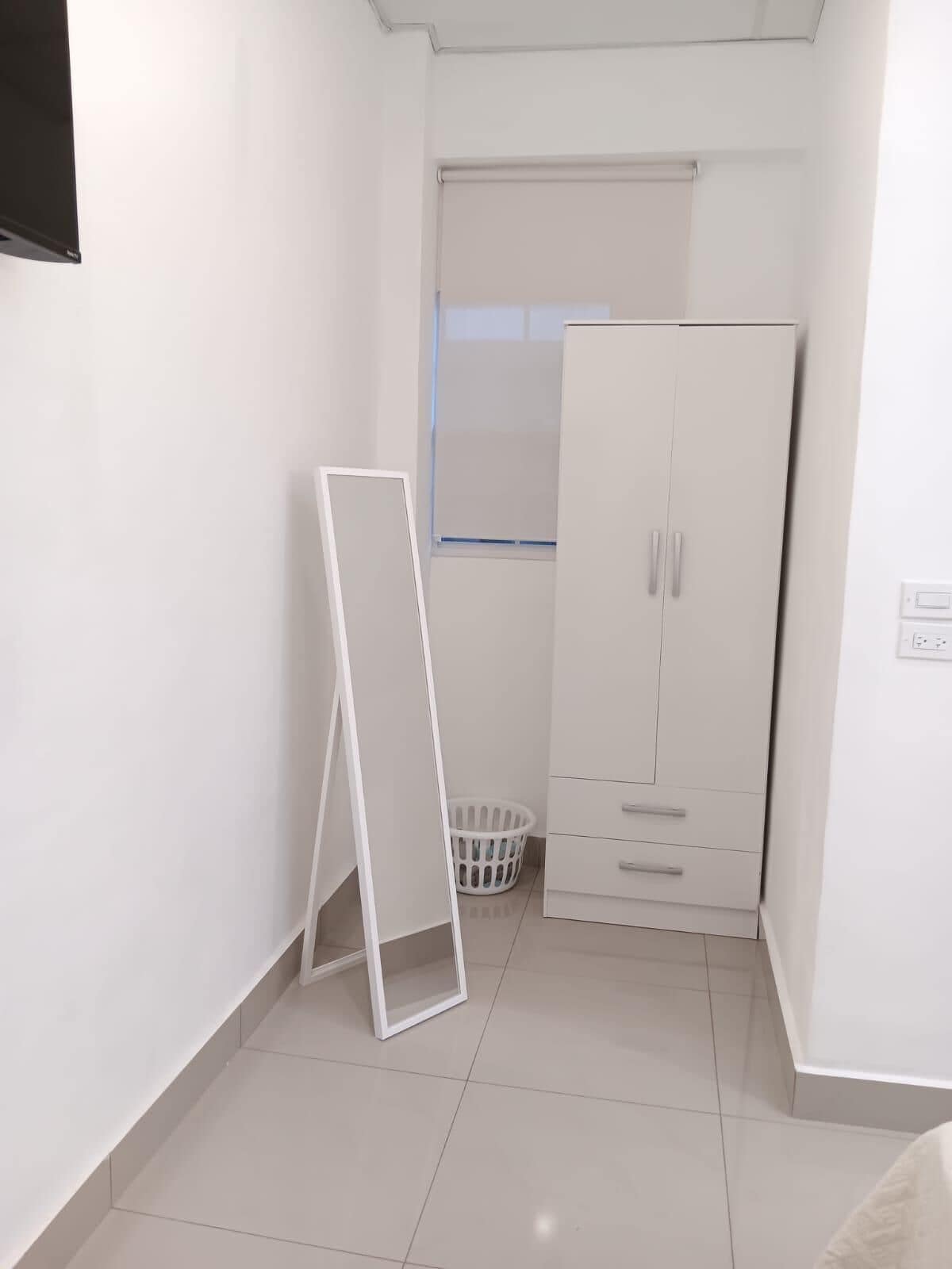 A small, white-walled room with a white wardrobe, a white laundry basket, and a white-framed easel.