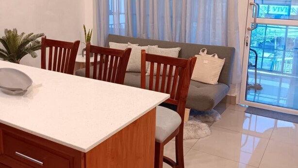 Una cocina moderna con armarios de madera y encimera blanca, un comedor con sillas de madera, un sofá gris y un ventanal con cortinas traslúcidas.